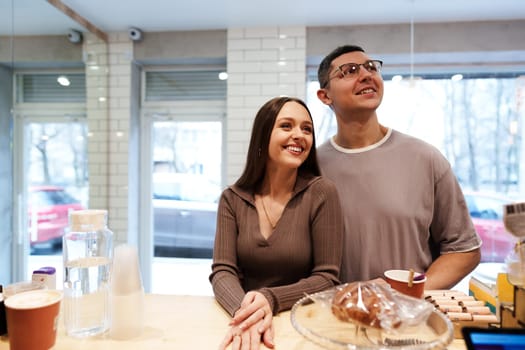 A couple stands together at a cafe counter, smiling and looking out the window. They are surrounded by coffee cups and baked goods, creating a warm and inviting atmosphere.