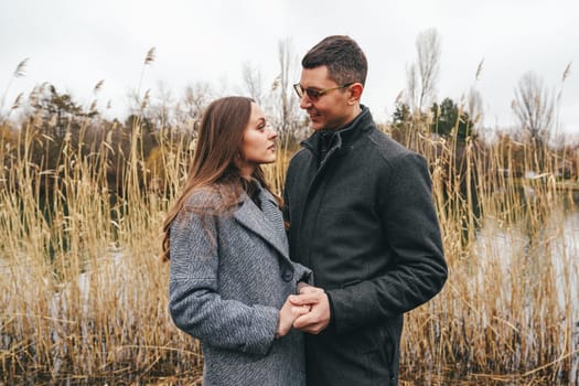 Two individuals share an intimate moment at the edge of a tranquil lake, surrounded by tall grass. The atmosphere is cozy with a hint of autumn chill.