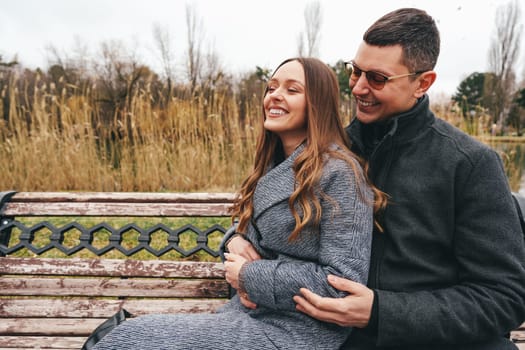 A couple sits closely on a park bench, laughing and sharing warm moments. Surrounding them are tall grasses and bare trees, depicting a cool day in nature.