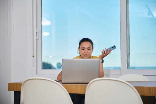 A professional woman manages tasks from her home office, utilizing a laptop and smartphone. The scene captures productivity and adaptability in a comfortable and modern space.