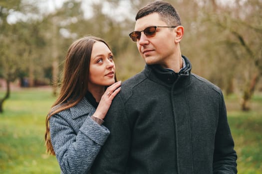 A couple stands close to each other in a lush green park. The woman gazes affectionately at the man, who is wearing sunglasses and a stylish coat.