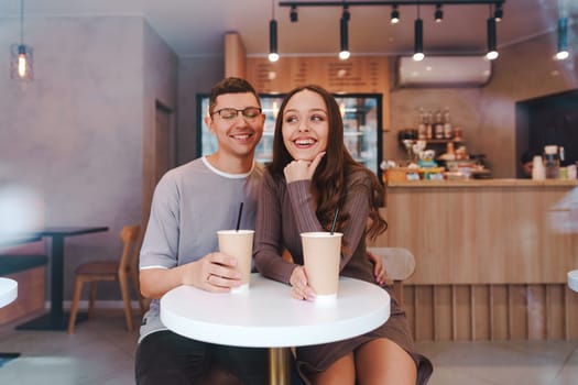 A young couple shares a joyful moment while sipping drinks at a modern cafe. They smile brightly as they engage in conversation at a small table.