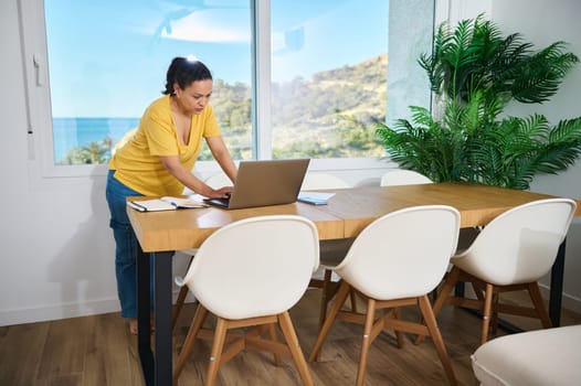 Professional woman working on a laptop in a stylish home office with a view of a serene coastal landscape, exemplifying focus and productivity in a comfortable environment.
