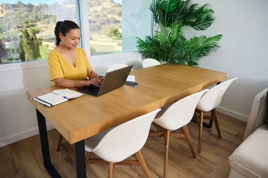 A confident woman working on a laptop in a stylish home office, emphasizing productivity, entrepreneurship, and a comfortable workspace, surrounded by a natural environment.