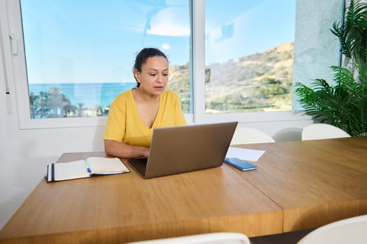 Confident woman working on her laptop in a bright home office with a scenic view outdoors. A peaceful combination of nature, productivity, and modern work lifestyle.