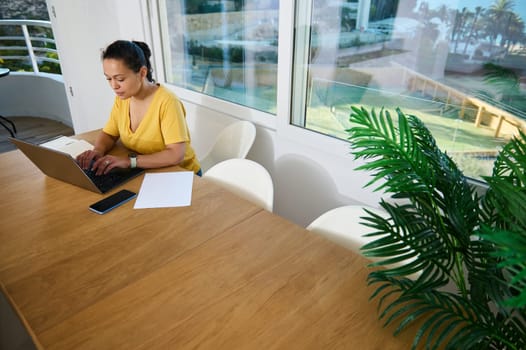 Woman working diligently on her laptop in a contemporary home office, surrounded by modern decor and a bright ambience, symbolizing productivity, comfort, and entrepreneurial spirit.