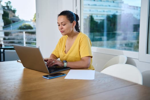 Focused woman in a yellow shirt working intently on her laptop while seated in a bright home office. Represents remote work, productivity, and entrepreneurship.