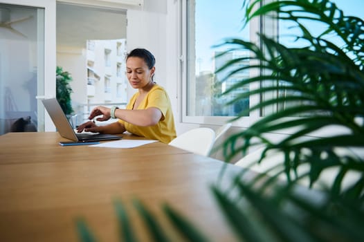 A confident woman in a bright home office working on her laptop surrounded by cozy decor, highlighting the modern remote work lifestyle. Her vibrant workspace conveys productivity and calmness.