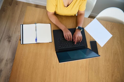 A woman in a yellow shirt types on a laptop at a wooden desk with a notebook and smartphone, combining efficiency and organization in a home office setup.