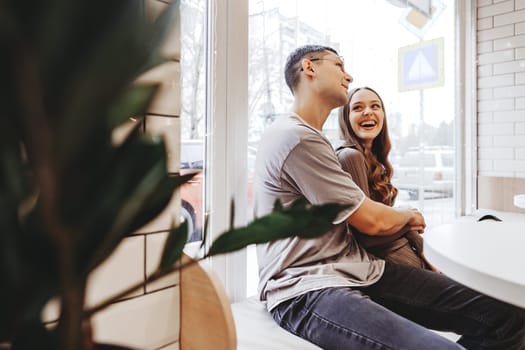 A couple sits closely at a table in a warm cafe, sharing smiles and laughter. Soft light filters through the windows, creating a relaxed atmosphere on a chilly day.
