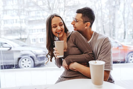 Two individuals share a joyful moment in a cozy cafe, drinking from paper cups. They are seated close together, surrounded by a wintery urban landscape visible through the window.