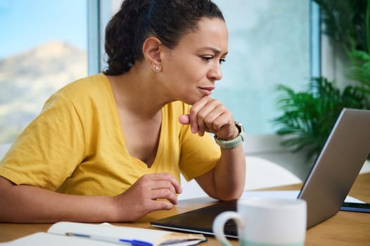 Woman in a yellow top focusing on work on her laptop. Background features a cozy home office. Concept of remote working, freelance, and comfortable productivity with modern devices.