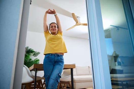 A cheerful woman stretches joyfully in her modern home interior, balancing work and leisure in her comfortable space, filled with natural accents and stylish decor.
