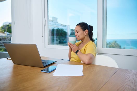 A woman in a bright home office enjoys a coffee break while working on her laptop, surrounded by a clean setup and beachside views. An inspiring blend of work and relaxation.