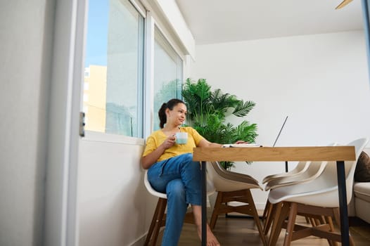 A woman enjoys a coffee break while working on her laptop in a bright, modern home interior. The setting conveys relaxation and productivity in a cozy and luminous workspace.