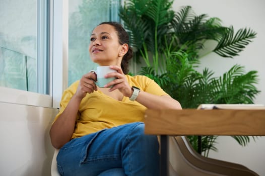 Woman enjoying a moment of relaxation while holding a coffee mug, surrounded by lush greenery and a cozy environment. Perfect representation of tranquility and comfort.
