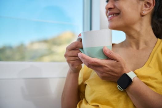 A cheerful woman enjoys coffee while relaxing near a window, embodying peace, comfort, and pleasant surroundings. The image highlights self-care moments and the serenity of a cozy environment.