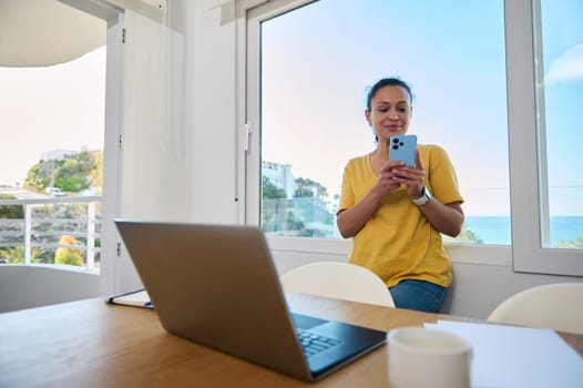 A confident woman in a bright office, working remotely with a laptop and smartphone, showcasing the digital nomad lifestyle and the balance between professionalism and outdoor inspiration.