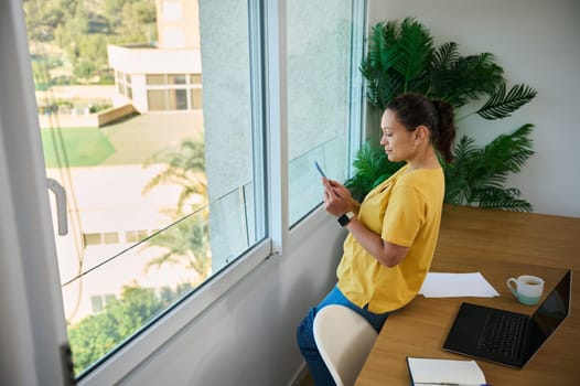 A business professional working at home, using a laptop and smartphone. The comfortable workspace features natural lighting, houseplants, and a tranquil atmosphere promoting productivity.