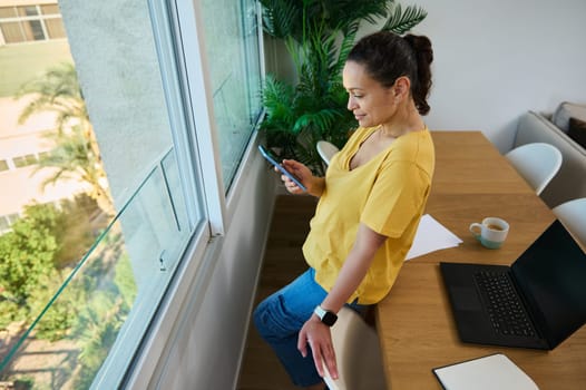 A woman leans by a window, using a smartphone during a break from work. She is in her home office setting with a laptop and coffee on the table nearby.