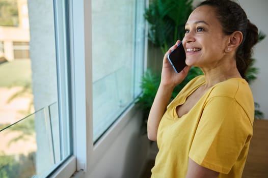 A joyful woman in a yellow top chats on her phone near a bright window. The setting emanates positivity and coziness, emphasizing connectivity and modern communication.