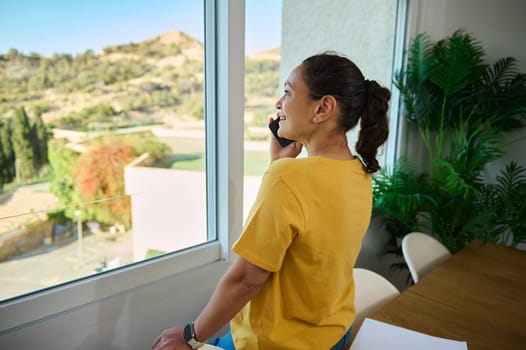 A cheerful woman stands by a window, smiling while on a phone call. The room features modern decor, lush plants, and an inspiring view of a sunny landscape, representing relaxation and connection.