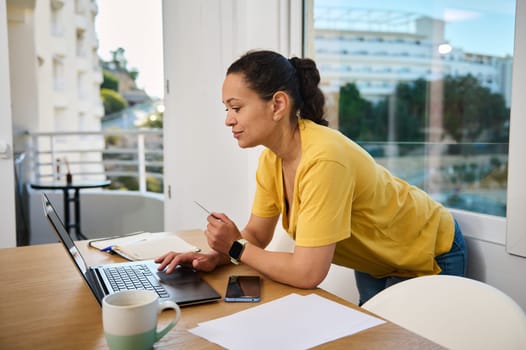 A woman in a yellow shirt works on a laptop at a desk in a bright indoor setting. A coffee cup, phone, and documents surround her, suggesting a productive working environment.