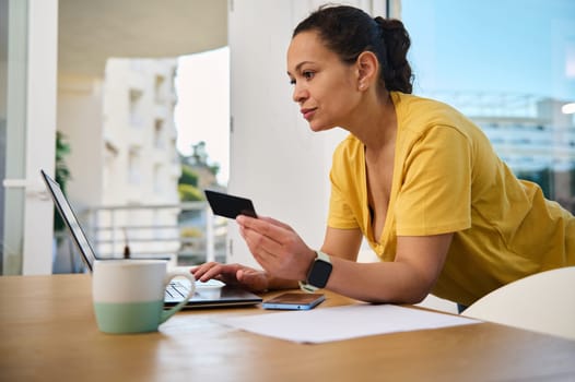 A woman in a yellow shirt works on her laptop from a home office, holding a credit card, examining information, and balancing work with a coffee break.