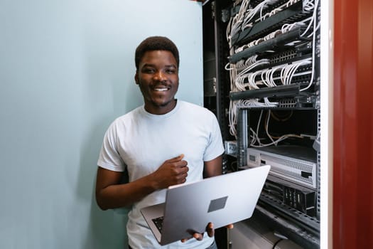 A technician is smiling while holding a laptop and standing in front of a server rack filled with network cables. The setting is bright and modern, indicating a data center environment.