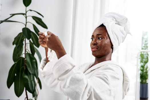 A woman wearing a white towel is applying a skincare treatment while looking in the mirror. The bathroom has bright lighting and green indoor plants, creating a refreshing atmosphere.