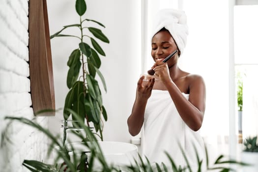 A woman enjoys her skincare routine, smiling as she applies a product in a well-lit bathroom adorned with greenery during the morning hours.