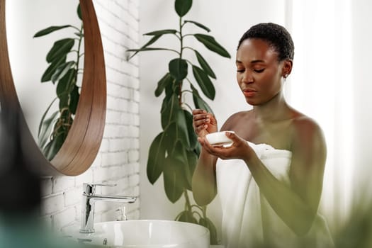 A young woman stands in a contemporary bathroom, wrapped in a towel, thoughtfully applying a skincare product. Natural light filters through the window, enhancing the serene atmosphere.