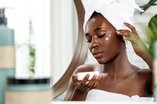 In a light-filled bathroom, a young woman gently applies moisturizer to her face while holding a small bowl in one hand. She appears relaxed and focused on her skincare ritual.