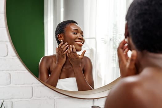 A woman with short hair smiles while applying skincare in front of a round mirror. The bright and clean bathroom features green accents and soft natural light, creating a serene atmosphere.