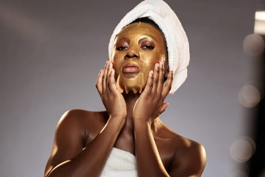 A young woman with a towel wrapped around her head gently applies a gold face mask to her skin during a beauty treatment session at a spa, exuding confidence and relaxation.