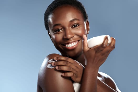 A smiling woman is applying moisturizer to her shoulder while holding a bowl of product. She enjoys her skincare routine, radiating happiness in a light blue setting.