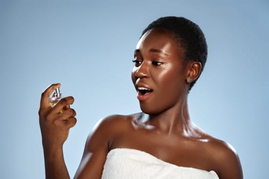 A woman with short hair expresses surprise and delight while holding a bottle of fragrance. She is wrapped in a towel against a light blue background in a studio during daylight.
