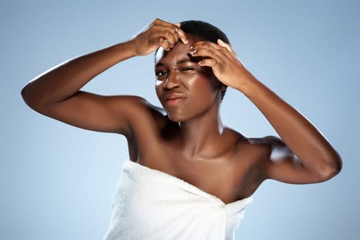 A woman with short hair is seen performing a skincare routine, targeting acne while wrapped in a towel. The backdrop is a soft, light blue, highlighting her expression of concentration.