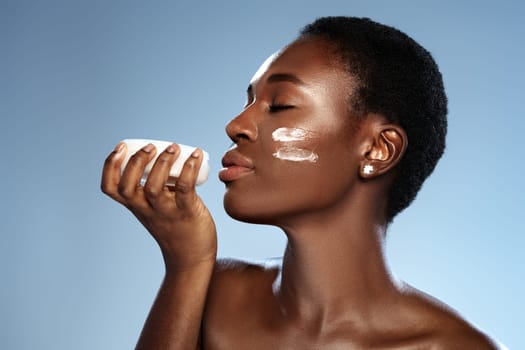 A woman holds a small bowl of skincare product close to her face while applying cream to her cheek. The bright blue background enhances a tranquil ambiance, emphasizing self-care.