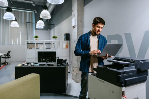 A young man stands by a printer in a modern office, working on his laptop. The space is bright and stylish, featuring contemporary decor and a relaxed atmosphere.