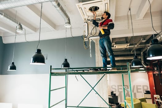 A man is working on an HVAC unit, using tools while standing on scaffolding in a modern office setting with industrial lighting and decor during daylight hours.