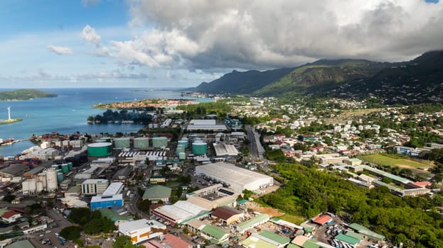 A coastal cityscape with turquoise waters, mountains, and green hills under a partly cloudy sky. Victoria city. Seychelles, Mahe.