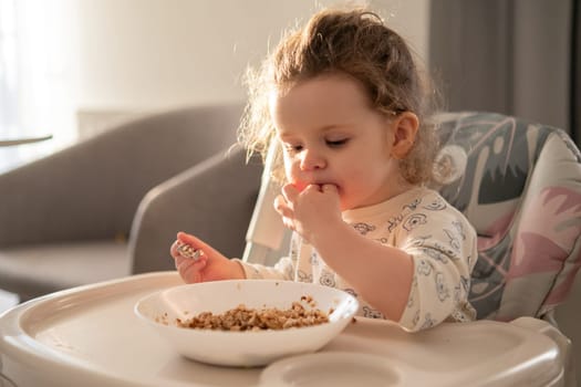 Curly haired toddler girl eating porridge in a high chair in a cozy kitchen. Morning routine and healthy breakfast time.