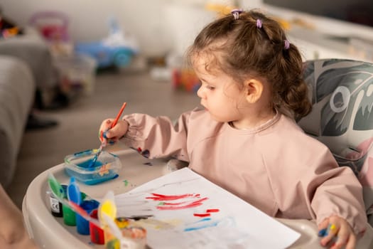 Cute toddler girl painting with gouache at a table near a sunlit window. Natural light highlights her artwork and the joyful atmosphere of creativity.
