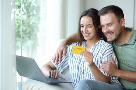 Happy couple buying online using card and laptop sitting in a couch in a house interior