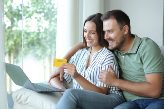 Happy couple buying online together with laptop and credit card sitting in a couch at home