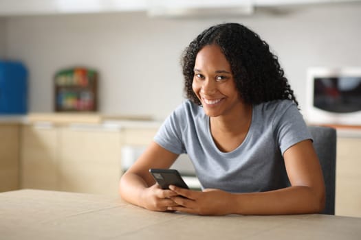 Happy black woman holding mobile phone looks at camera in the kitchen at home