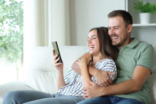 Happy couple enjoying watching media on mobile phone sitting on a couch at home