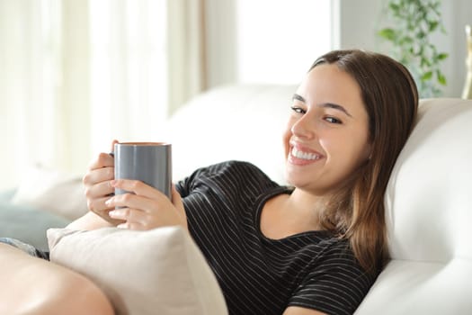 Portrait of a happy woman holding coffee cup smiling sitting in a couch at home
