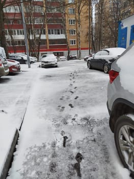 A narrow sidewalk has footprints through the fresh snow. Several cars are parked nearby, some with snow on top. An apartment building is visible in the background.